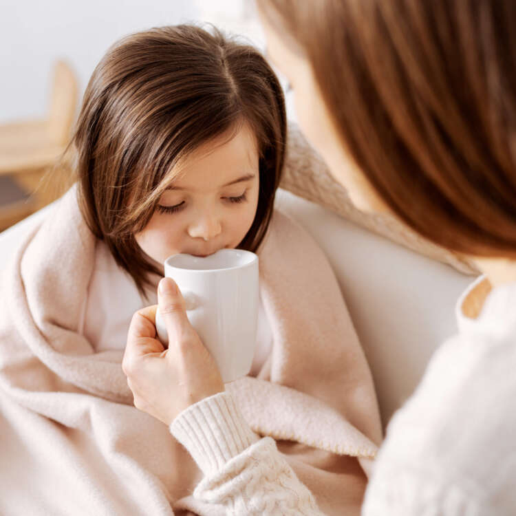 Ein Kind sitzt eingewickelt in eine Decke auf dem Sofa und bekommt von seiner Mutter eine Tasse an den Mund geführt