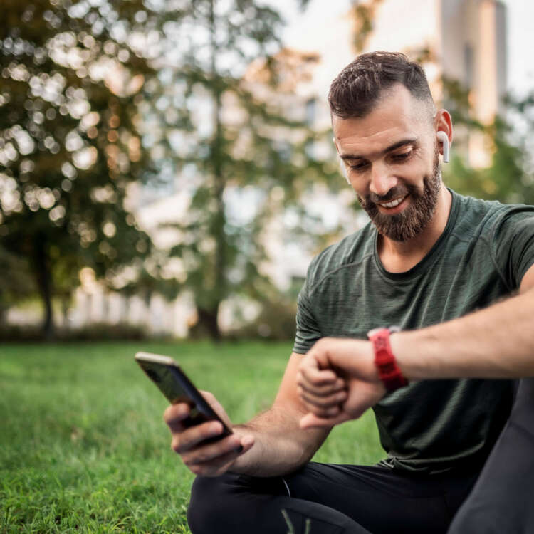 Ein Mann sitzt in einem Park mit einem Handy in der rechten Hand, er schaut auf die Uhr an seinem linken Arm