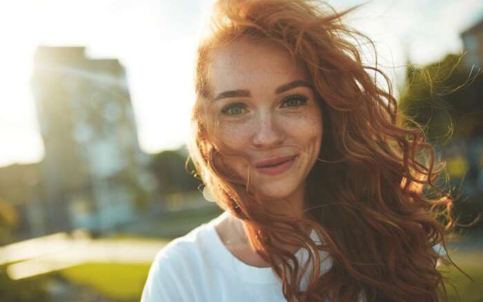 Eine rothaarige Frau mit Sommersprossen lächelt in die Kamera, die Haare sind vom Winde verweht