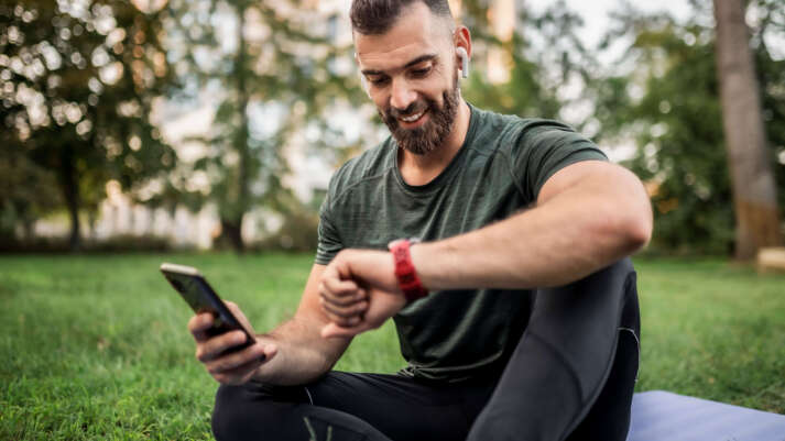 Ein Mann sitzt in einem Park mit einem Handy in der rechten Hand, er schaut auf die Uhr an seinem linken Arm