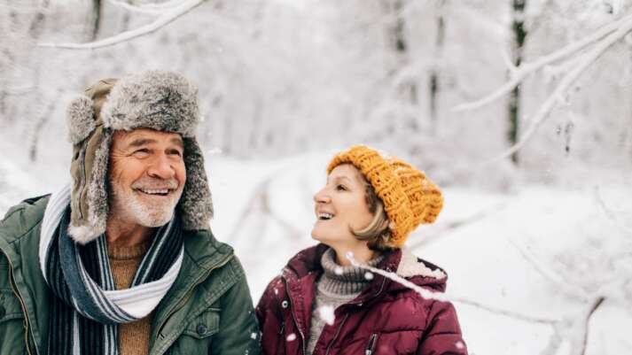 Ein lächelndes Paar warm eingepackt mit Windermütze und -Jacke steht inmitten einer Schnee-Landschaft