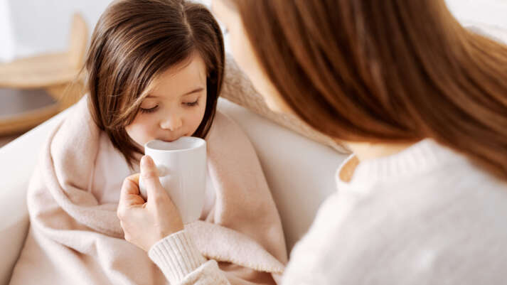 Ein Kind sitzt eingewickelt in eine Decke auf dem Sofa und bekommt von seiner Mutter eine Tasse an den Mund geführt