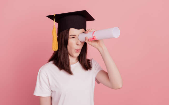 Eine junge Person mit Absolventenhut nutzt ihr Diplom als Fernrohr